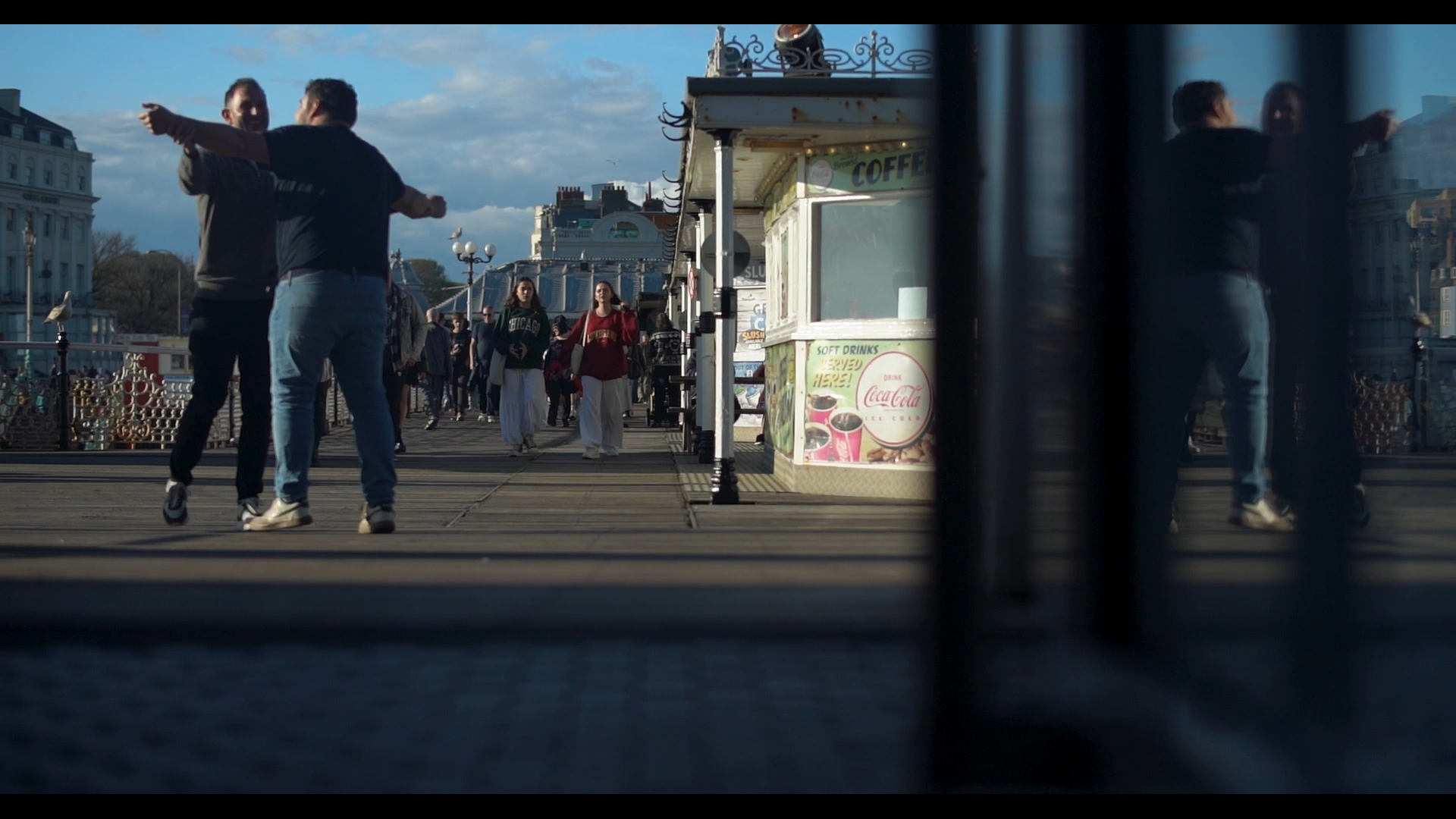 Two men dancing or embracing on a pier, with other people walking in the background and shops along the pier, some of which sell drinks and snacks.