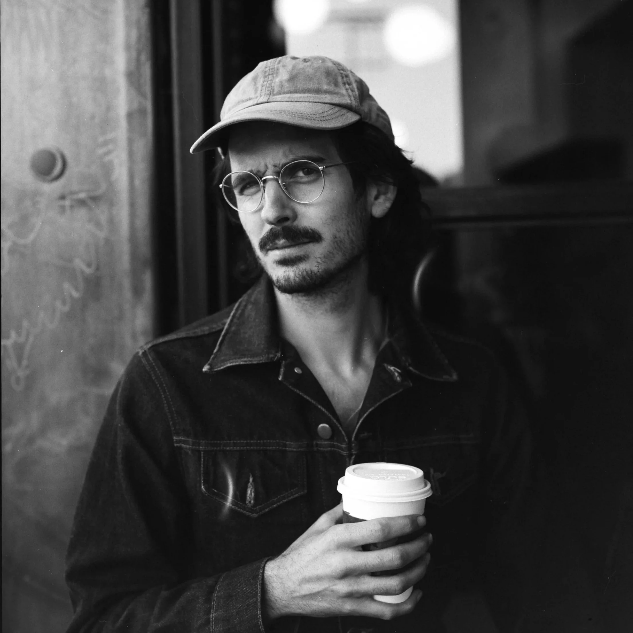 A man with glasses and a mustache wearing a denim jacket and a cap, holding a coffee cup, sitting indoors.