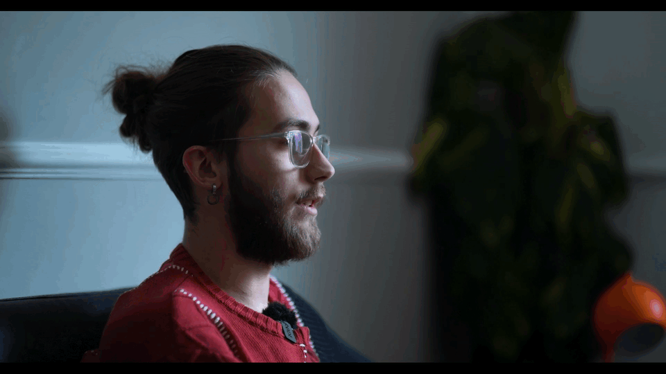 Profile of a man with glasses and a beard, sitting indoors, with a plant and a lamp in the background.