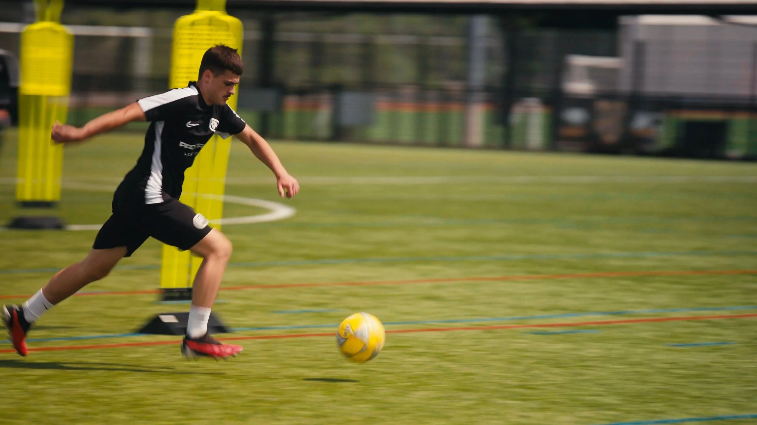 A young male soccer player in black uniform running on a soccer field during practice or game, chasing a yellow soccer ball.