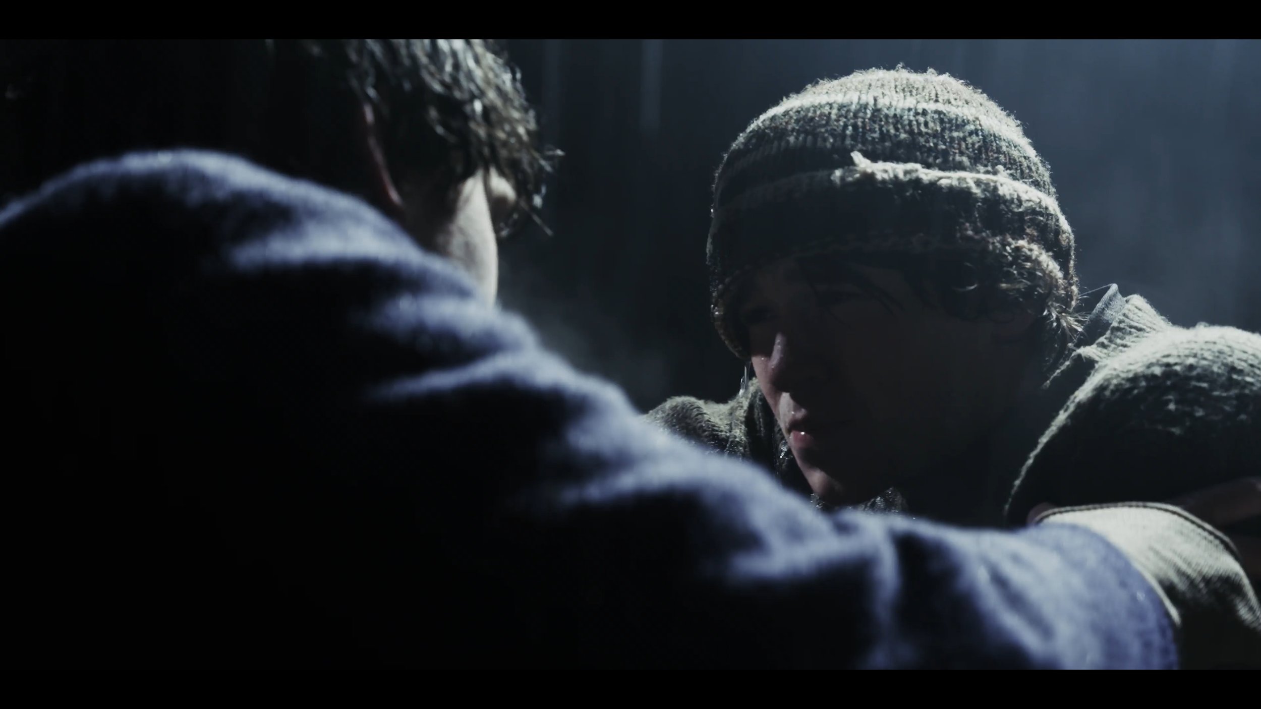 Two young men with dark hair face each other closely, one wearing a beanie, in a dimly lit scene.