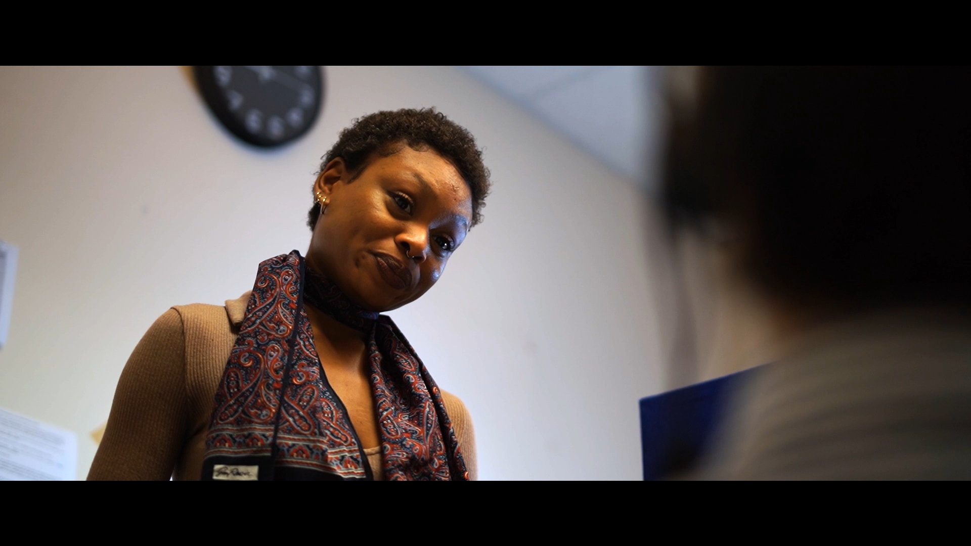 A woman with short curly hair, wearing gold earrings, a patterned scarf, and a tan sweater, looking down at a blue object, with a clock on the wall in the background.