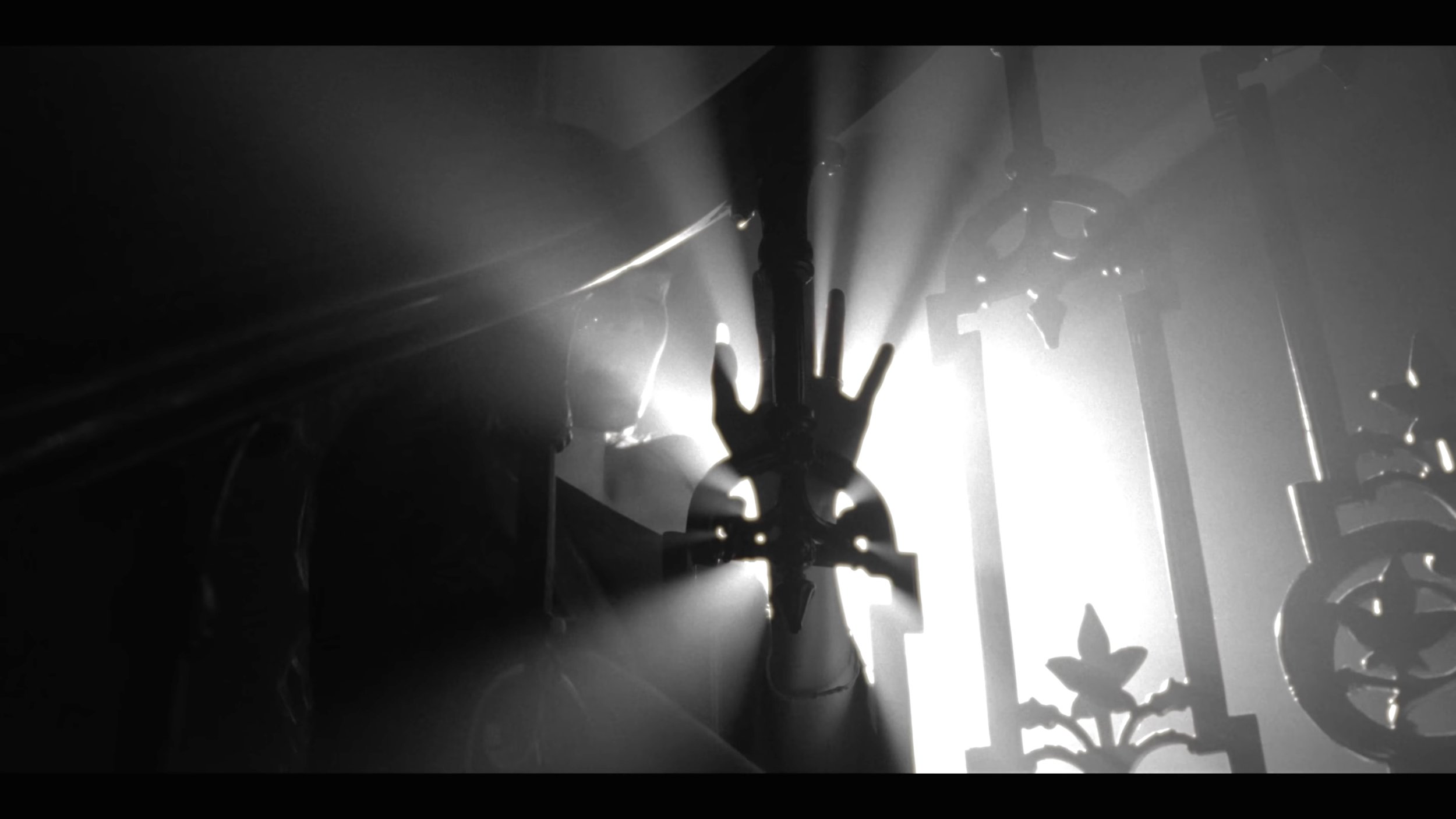 Black and white photo of a hand reaching out and touching a metal structure with decorative elements, backlit creating a silhouette and light rays.