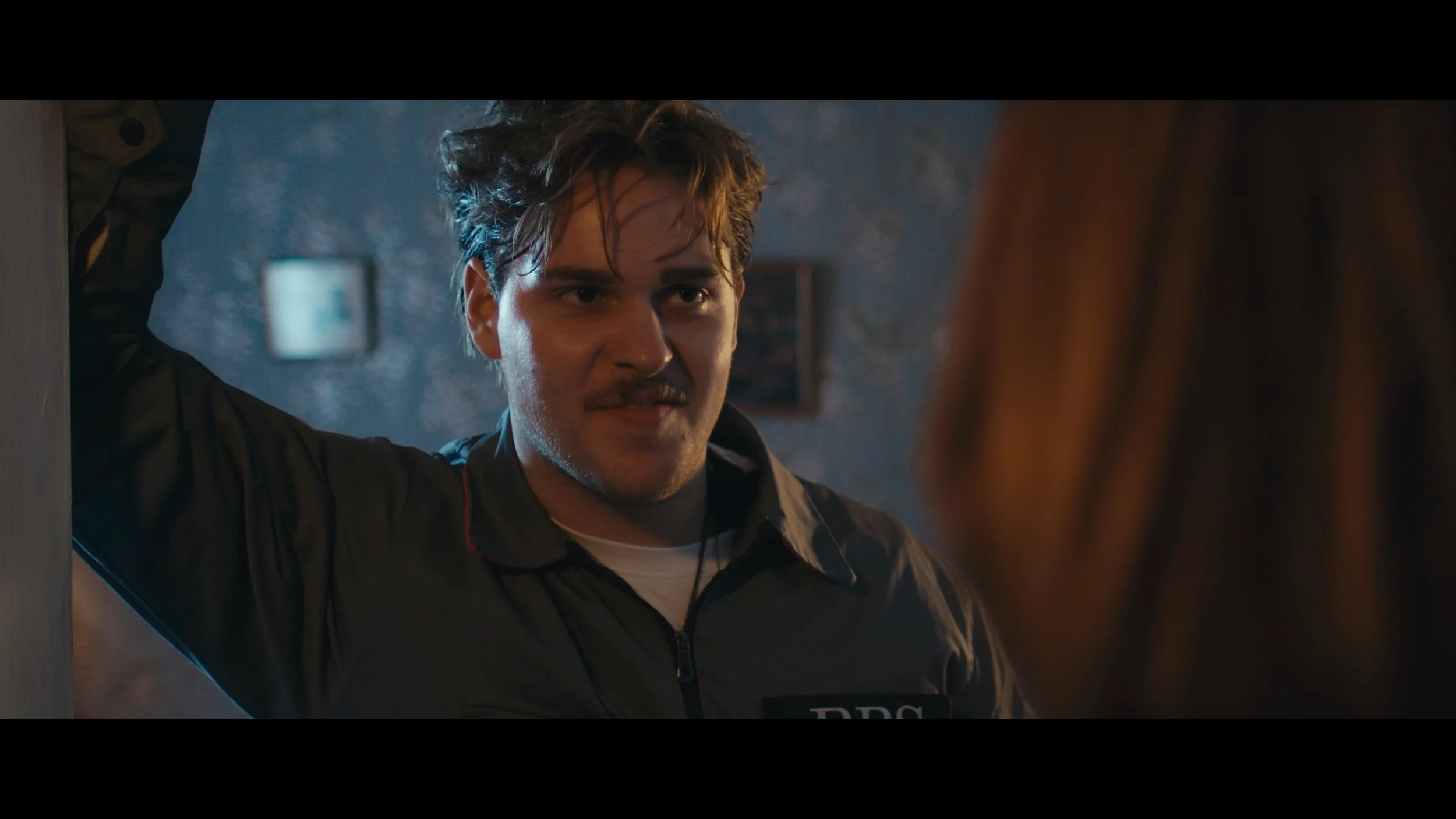 A young man with messy hair, wearing a black jacket with a badge labeled 'RPC', is standing in a dimly lit room and looking intensely at a person with long reddish-brown hair.