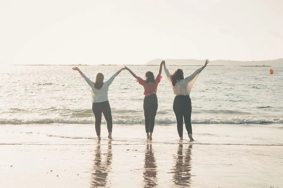 Three woman at the beach with hands raised in the air. How life could feel with Blossom Within Online Counselling