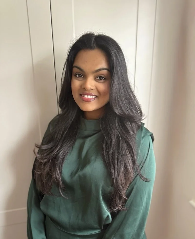 Annie Khedia, qualified therapist of Blossom Within. She has long, wavy black hair wearing a green blouse, smiling and standing indoors in front of a light-colored wall.