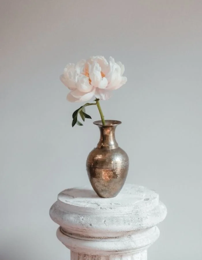 Blossom Within pink peony flower in a small copper vase on a white pedestal