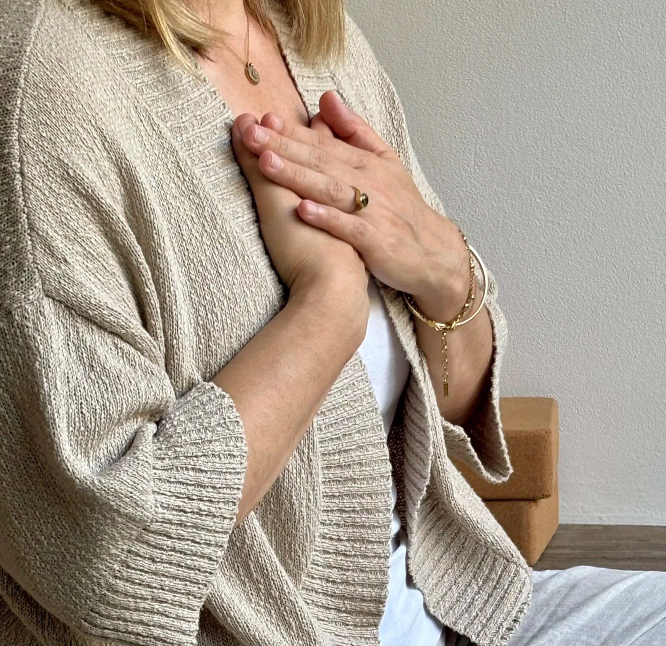 A woman with blonde hair is placing her hands over her chest in a gesture of prayer or gratitude. She is wearing a beige knitted cardigan, jewelry including a necklace, rings, and bracelets, and a white top.