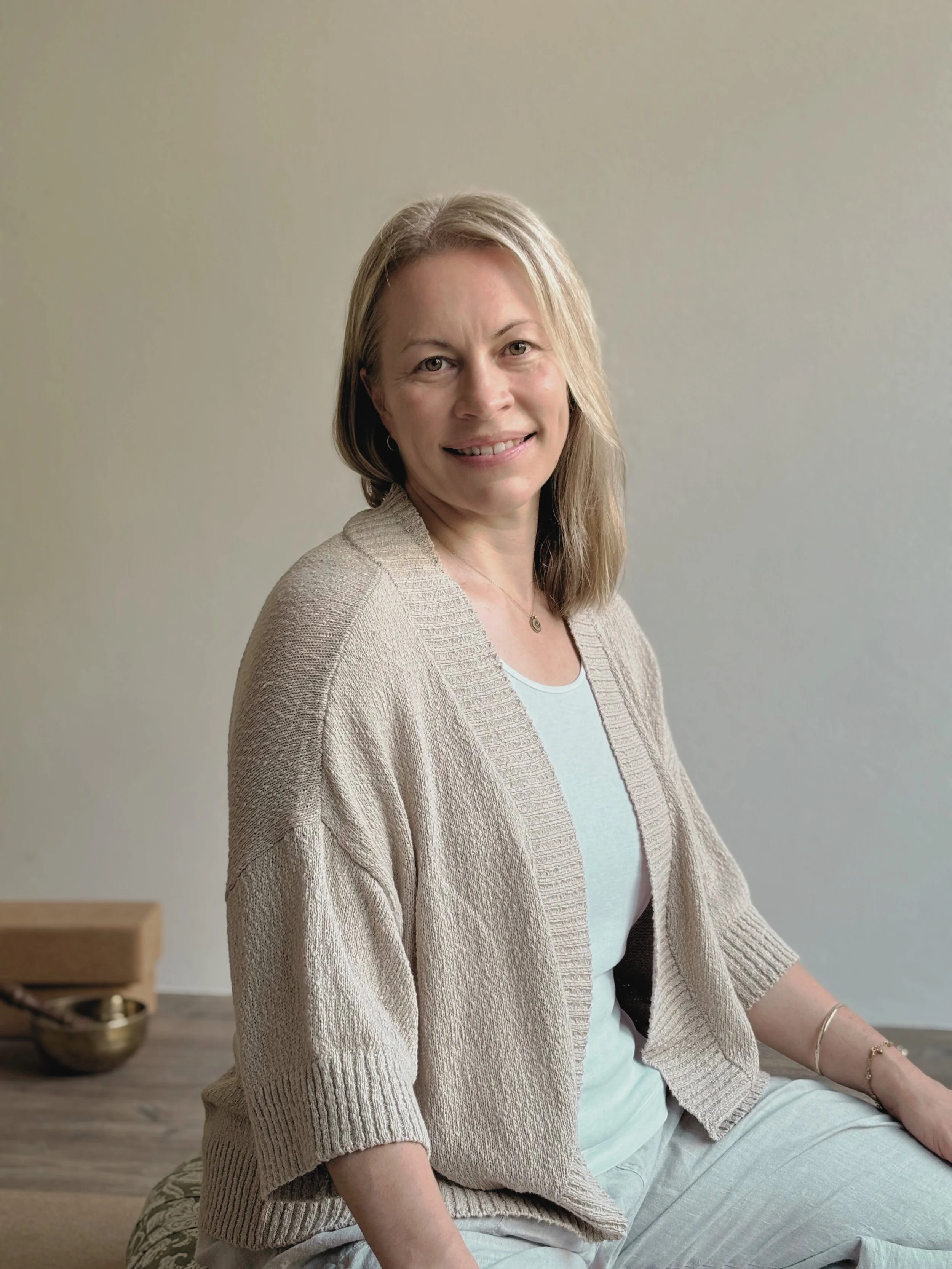 A woman with blonde hair wearing a beige cardigan and a white shirt, sitting indoors with a light-colored wall behind her.