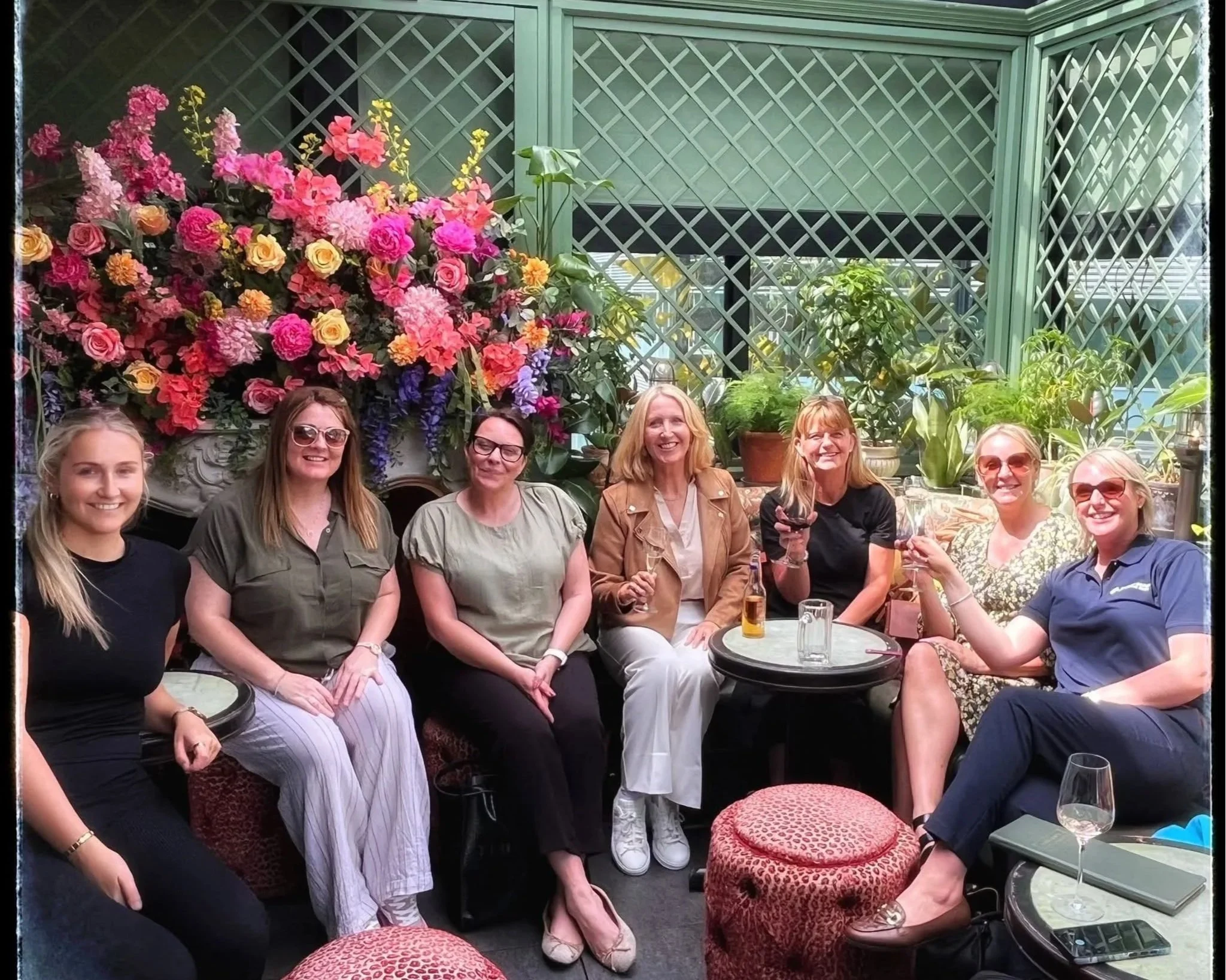 Group of women sitting together at a table in an indoor garden or greenhouse surrounded by colorful flowers and green plants, smiling and enjoying each other's company.