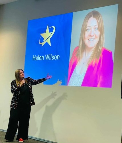 A woman is giving a presentation in front of a large screen displaying her name, Helen Wilson, and her photo with a blue logo featuring a star and a swoosh.