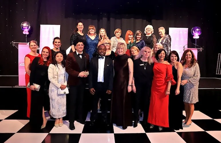 Group of people in formal attire at an event with a black and white checkered floor and a stage with three pink banners and balloons in the background.