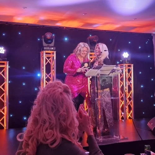 Two women on stage at an event, smiling and looking at papers, with a woman in the foreground clapping. The stage has a dark backdrop with small blue lights and orange light trusses, and an orange and purple sunset ceiling.