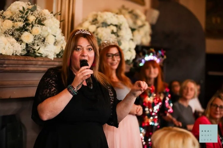 A woman in a black dress with a tiara speaking into a microphone at a celebration, with other women and floral decorations in the background.