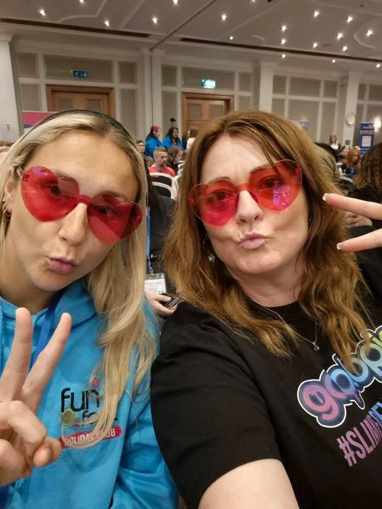 Helen Wilson and another woman wearing red heart-shaped sunglasses taking a selfie indoors at a crowded event or conference, making peace signs and posing playfully.