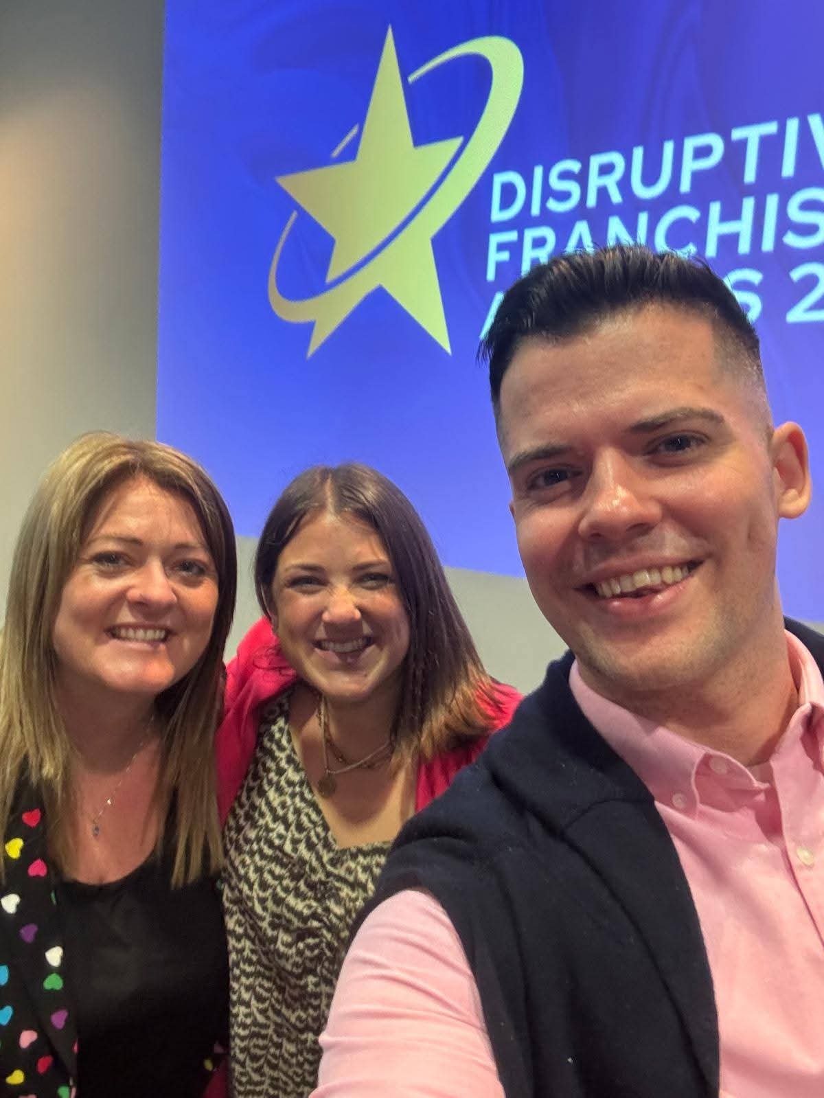 Three smiling people taking a selfie in front of a blue presentation slide with a star logo and the text 'Disruptive Franchising 2023'