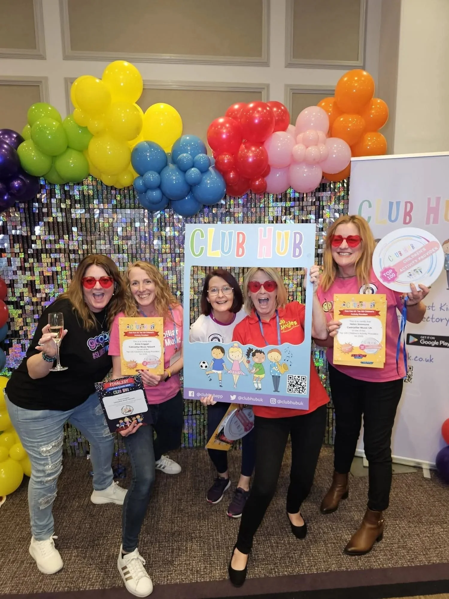 A group of five women celebrating at an event, holding signs and certificates, with colorful balloons and a sparkly backdrop in the background.