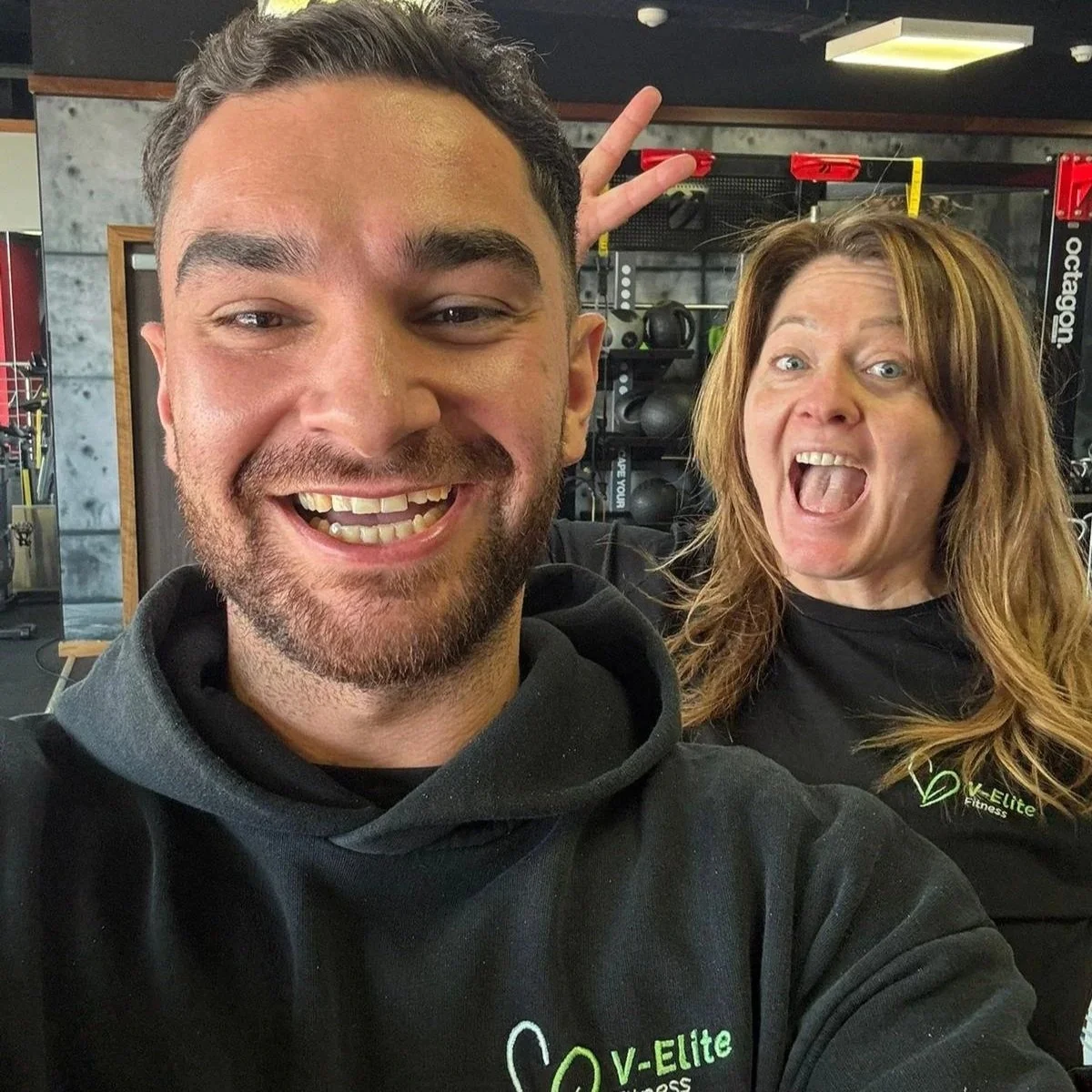 Helen and her PT taking a selfie in a gym, smiling and making peace signs, with gym equipment in the background.