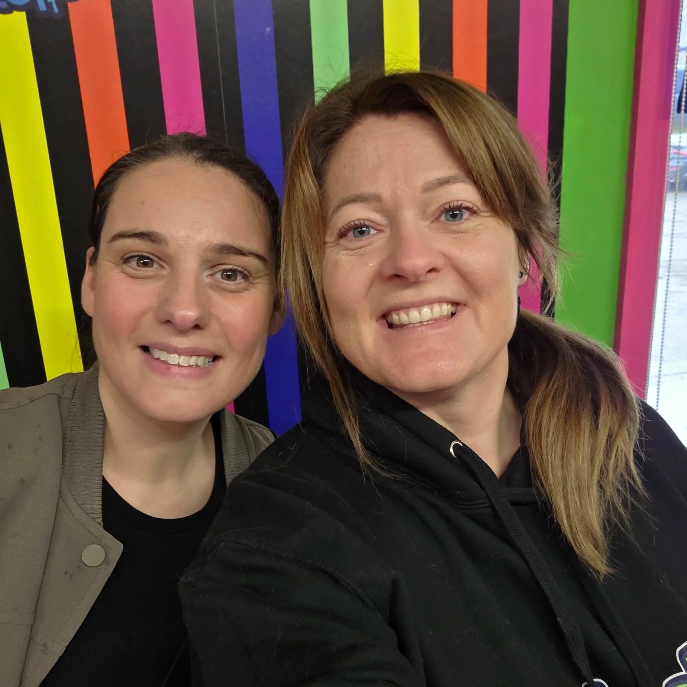 Helen and Rachel smiling, standing close together in front of a colourful background with rainbow-coloured vertical stripes.