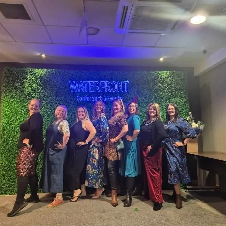 Group of nine women standing in front of a green wall with a blue neon sign that reads 'WATERFRONT Conference & Events,' posing with smiles and different outfits.