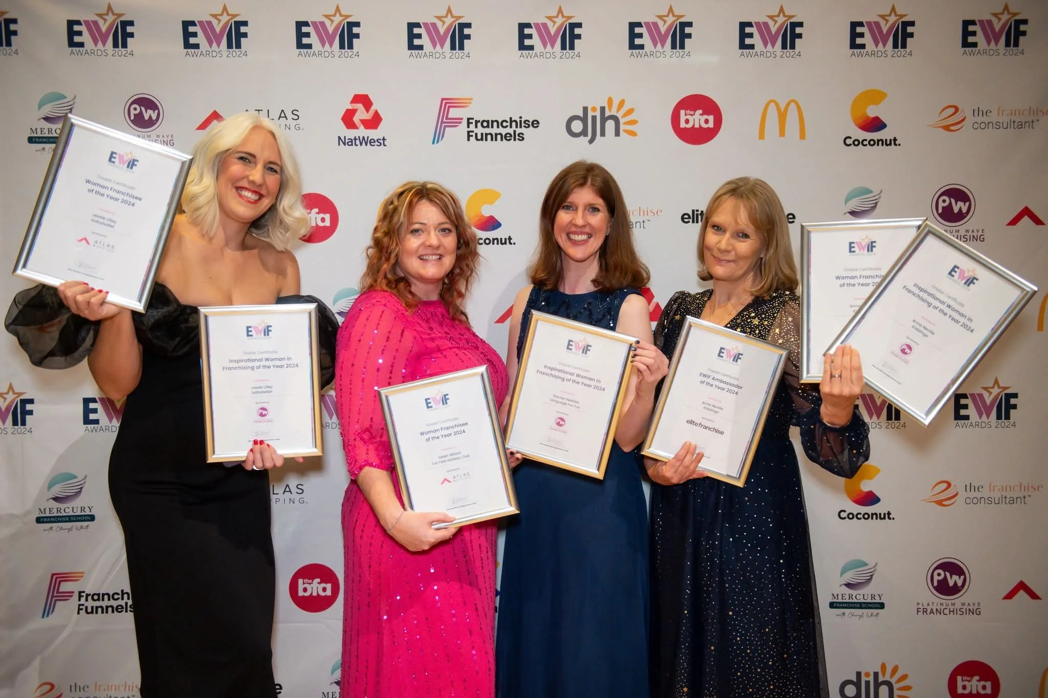 A woman wearing a tiara and black dress speaking into a microphone at an indoor event with floral decorations and a mirror in the background.