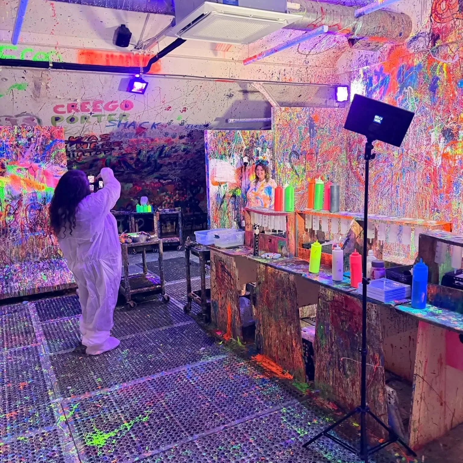 Indoor paint splatter art studio with splattered walls, floor, and furniture. Two people are present: one person taking a photo, and another model standing against the splattered wall. Bright graffiti-style words on the wall, colorful paint bottles on the counters, and studio lighting equipment.