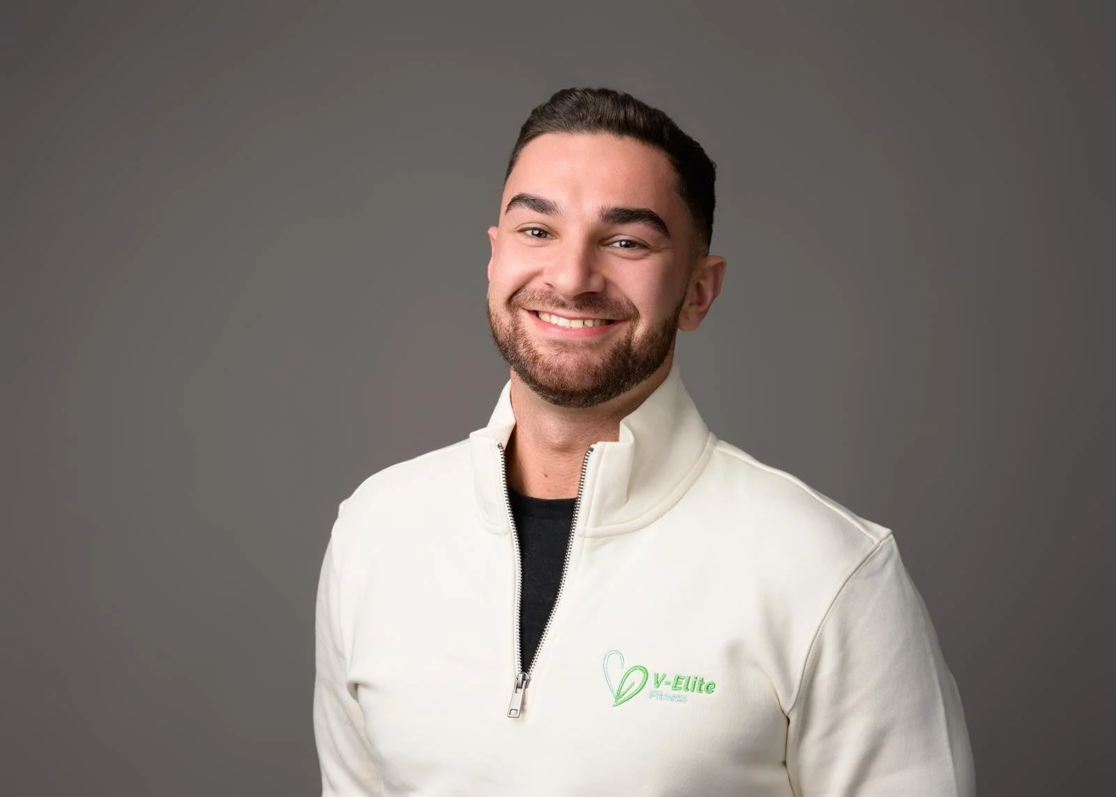 Vedat Dogan, PT, wearing a white jacket with a zipper and a logo that reads "V-Elite" on the left side, standing against a neutral grey background.