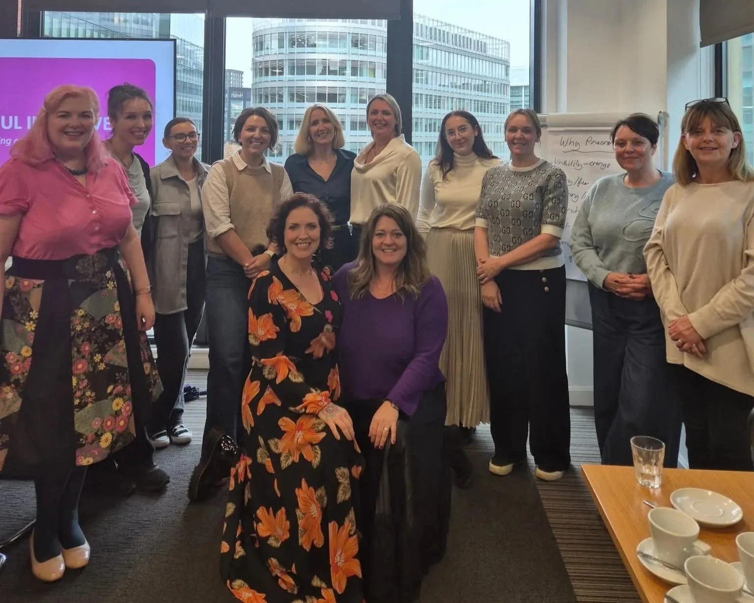 A group of women gathered in an office with large windows overlooking city buildings, posing for a photo during a meeting or workshop.
