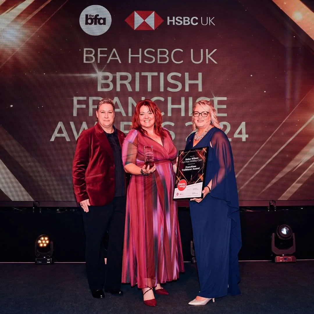 Three women in formal dresses on stage, one receiving an award from another, with a third woman smiling and clapping.