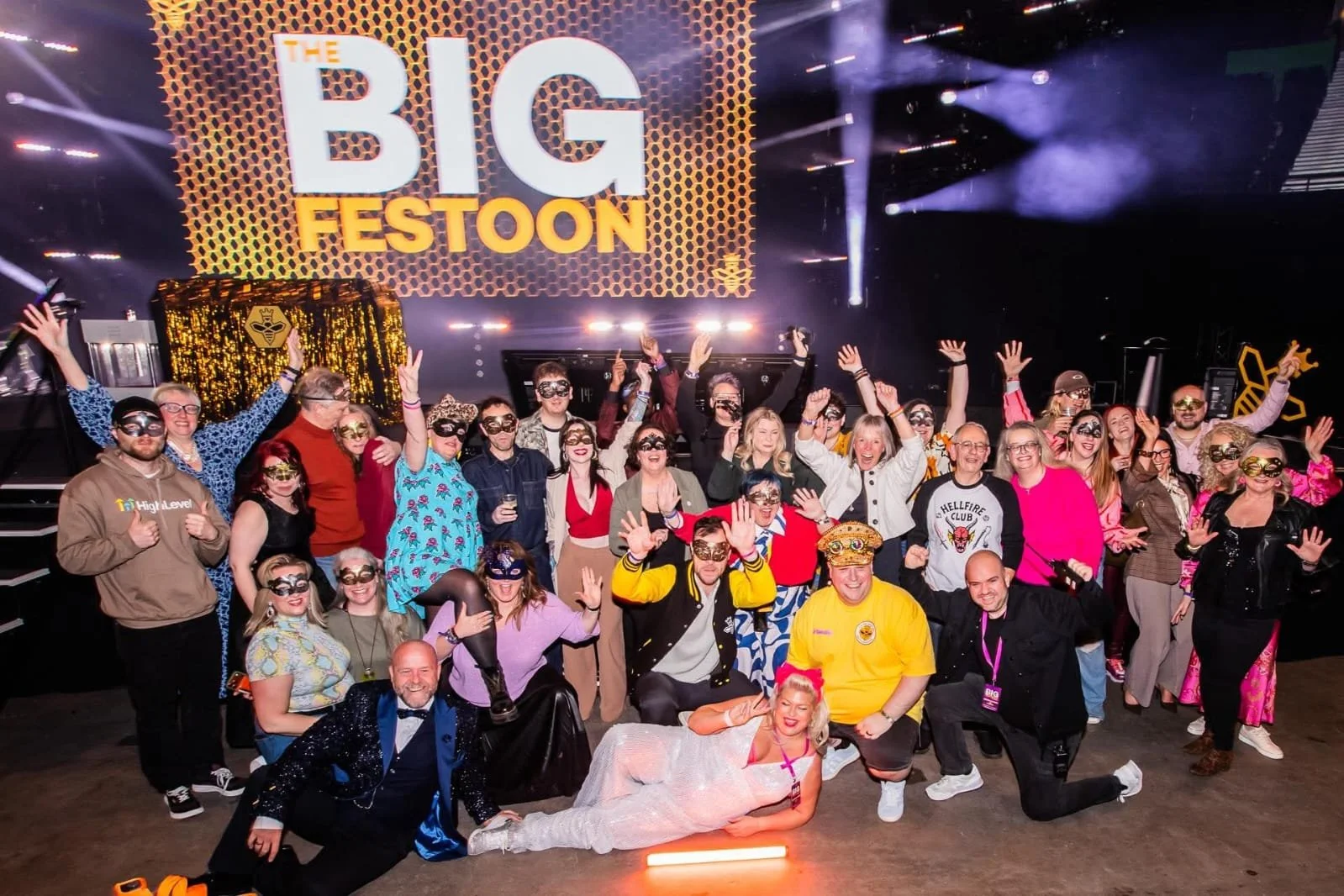A group of people celebrating at a festival with a large sign that reads 'The Big Festoon' in the background. Many are wearing masks, costumes, and colorful clothing, with some raising their hands and smiling for the camera. There is a woman lying on the ground in a sparkly dress, and a man in a yellow shirt and a woman in a blue jacket are prominent in the front.
