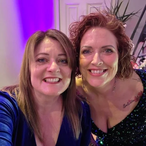 Helen Wilson and Susie Sprigg smiling for a selfie at a celebration or party, with purple and white lighting in the background.
