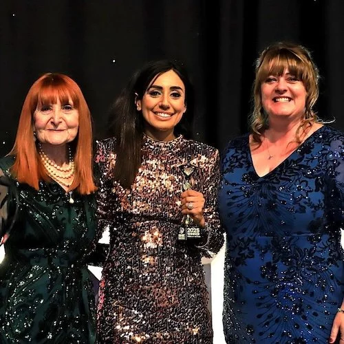 Sue France, Helen Wilson and the award winner standing together, smiling at a formal event against a black backdrop.