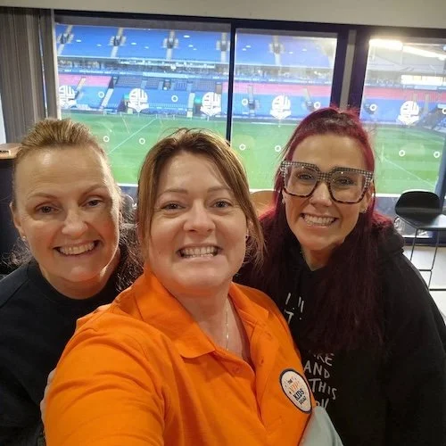 Three business women smiling in front of a large window overlooking a sports stadium.