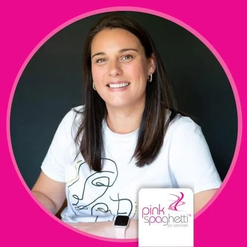 A woman with long dark hair smiling at the camera, wearing a white T-shirt with a line drawing of a face. She is sitting at a table with a smartwatch on her wrist. The background is dark gray with a pink circular border. There is a Pink Spaghetti logo and sign on the table.