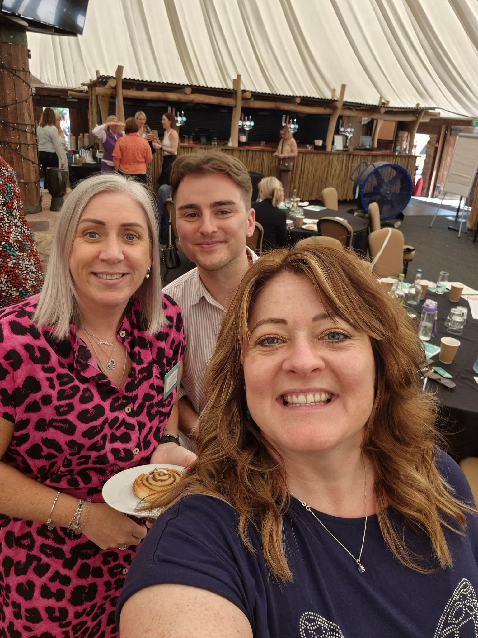 Selfie of four women smiling in a social setting with a bar and other people in the background. One woman is holding a plate with a cinnamon roll.