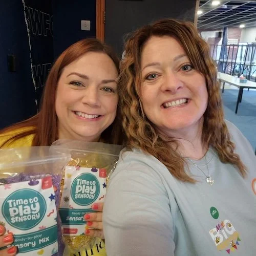 Two women smiling, holding bags of sensory popcorn, standing indoors in a well-lit space with tables in the background.