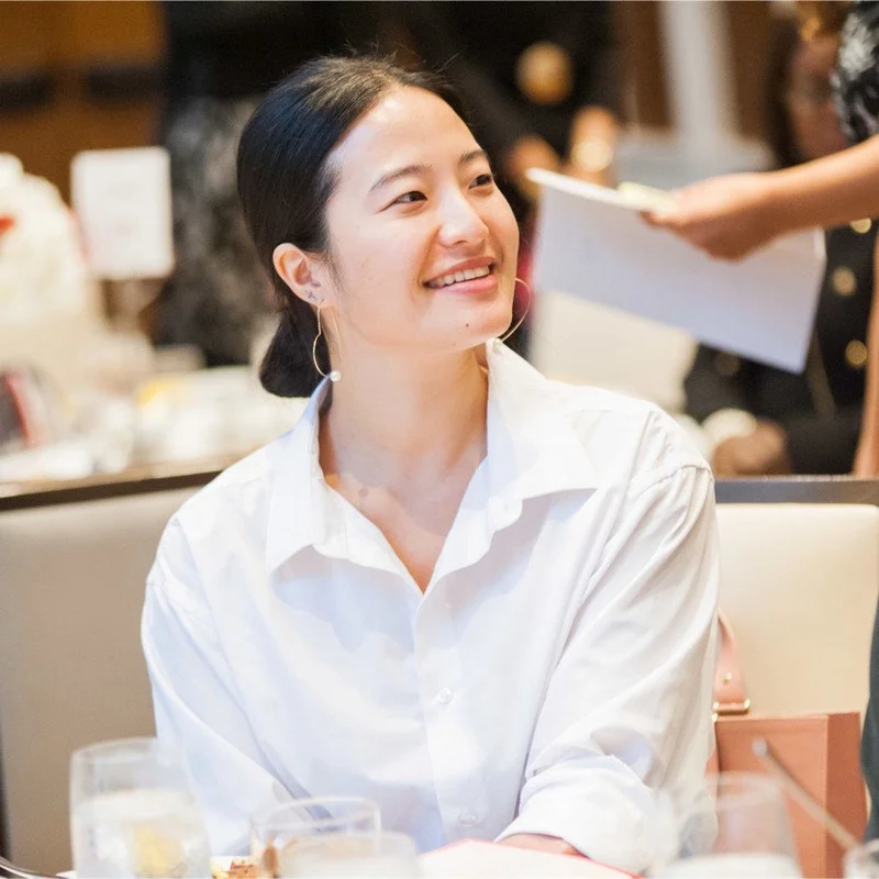 A woman in a white shirt smiling, seated at a table in a restaurant or cafe.