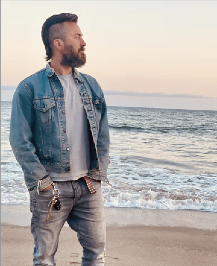 A man with a beard and styled hair stands on the beach near the shoreline, looking to the left, wearing a denim jacket over a gray shirt, with keys hanging from his pocket during sunset.