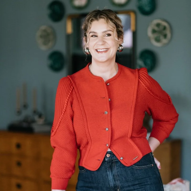 A young woman with short curly hair and earrings, smiling, wearing a red cardigan and dark jeans, standing indoors with a blurred background of wall decorations and a wooden dresser.