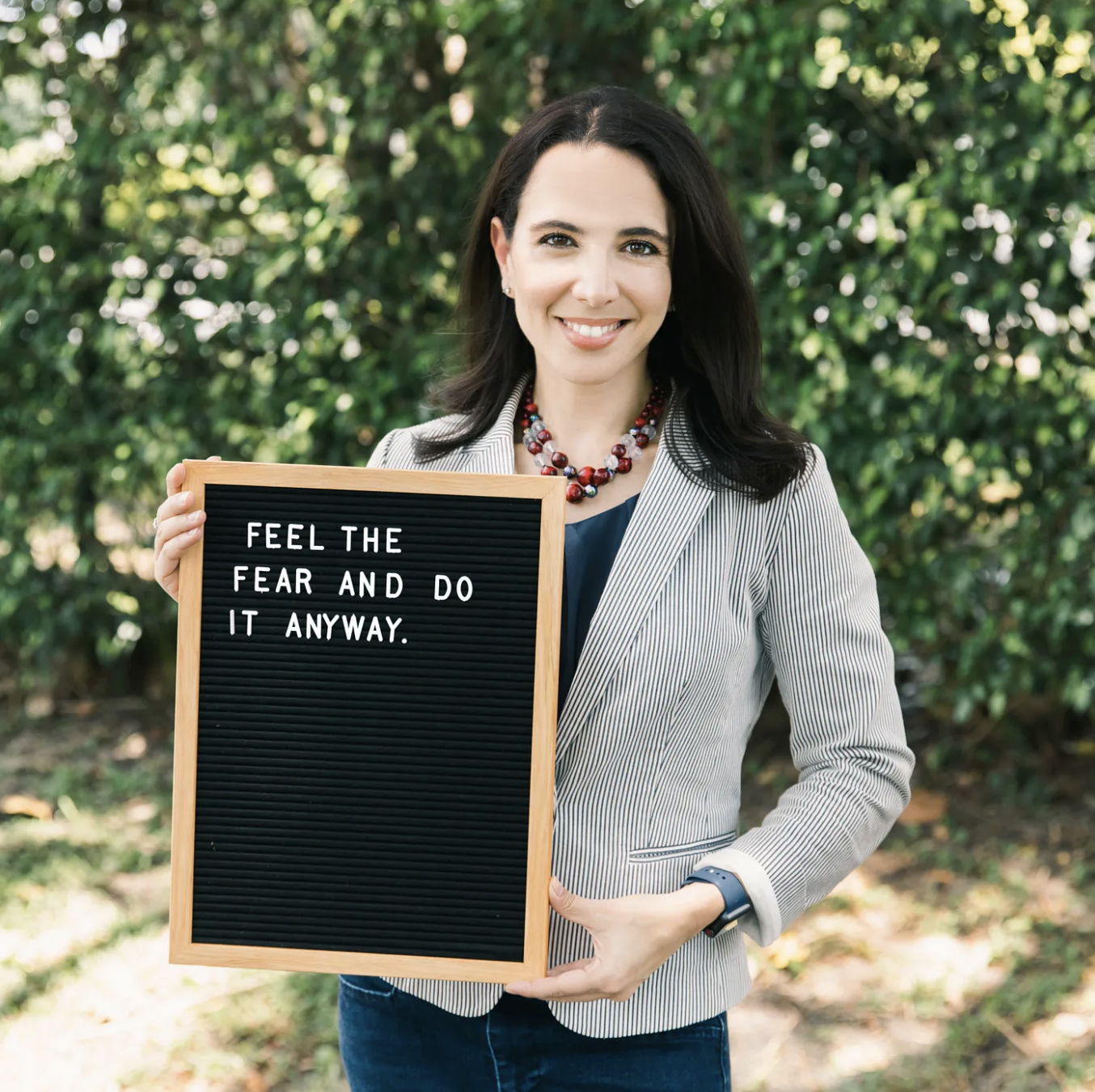 A woman smiling outdoors holding a black message board that reads, 'Feel the fear and do it anyway.' She is wearing a light striped blazer, a navy shirt, a colorful beaded necklace, and a smartwatch, with a background of green foliage.