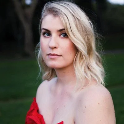 Portrait of a young woman with blonde hair, wearing a red top, standing outdoors in a park with green grass and trees in the background.