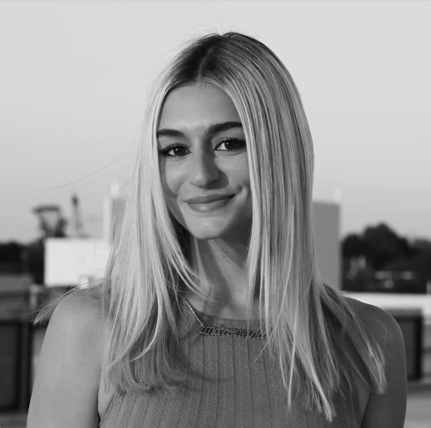 Black and white photo of a young woman with long blonde hair, smiling, wearing a sleeveless top and a necklace that says "Madonna" on a rooftop.