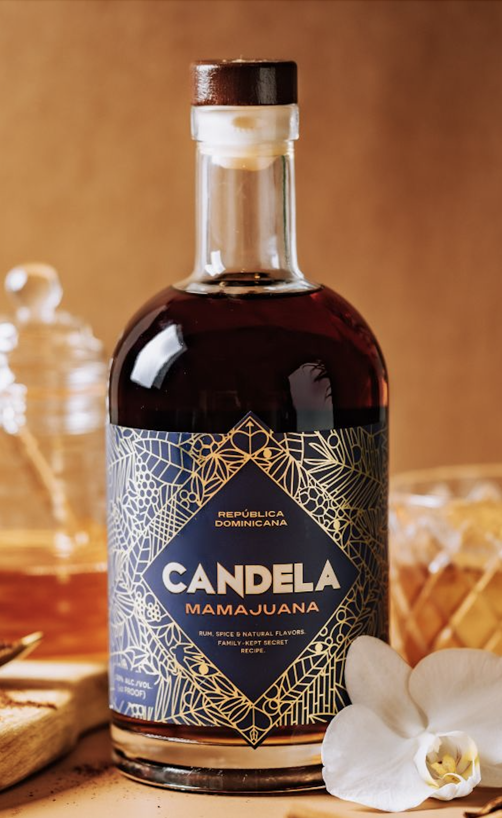 A bottle of Candela Mamajuana, a Dominican Republic herbal liquor with a dark amber color, on a wooden surface with a white orchid flower in front.