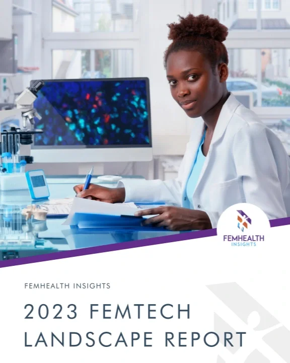 A woman scientist in a white lab coat sitting at a lab bench with a notebook and pen, in a laboratory with standard equipment and a computer monitor in the background.