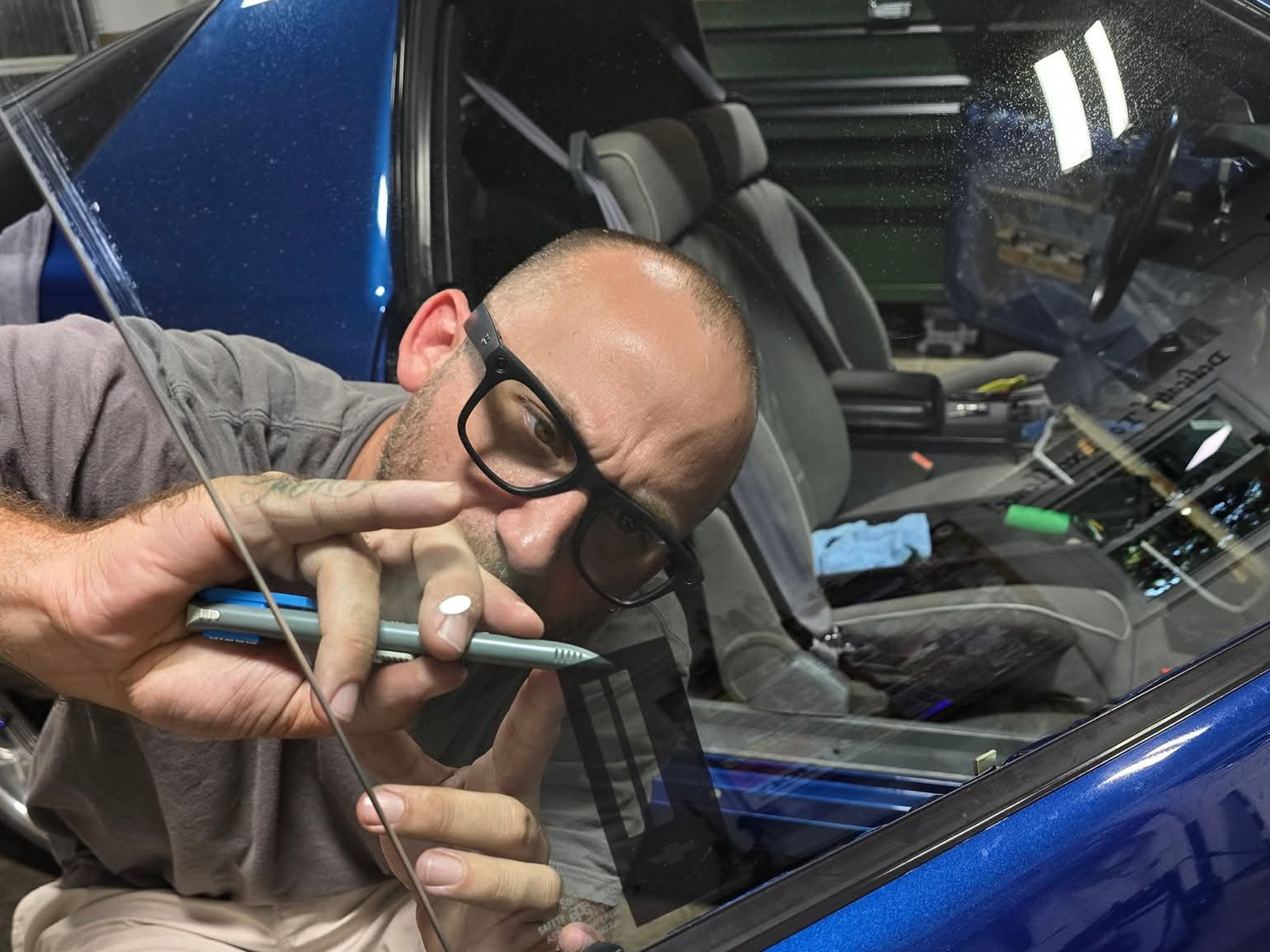 A man with glasses taking a selfie through the window of a blue car in a garage or workshop.