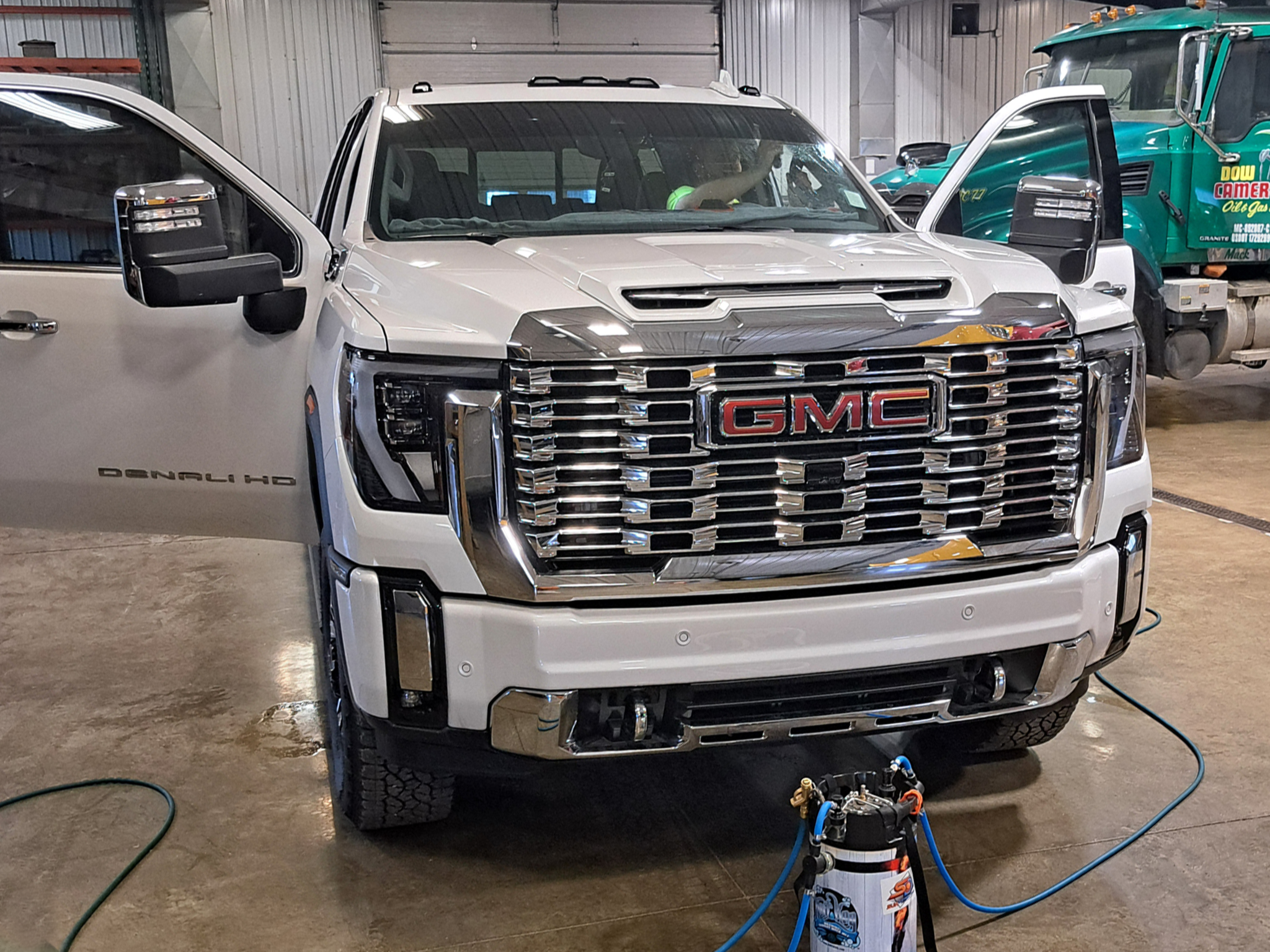White GMC Denali HD pickup truck inside a workshop with open driver side door, connected to testing equipment on the floor, with a green truck and workshop interior visible in the background.