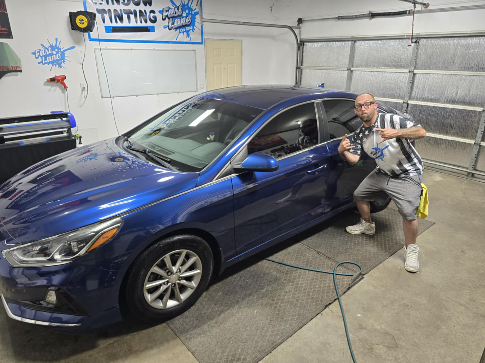 A man posing next to a blue sedan inside a car wash facility with signs and equipment on the walls.
