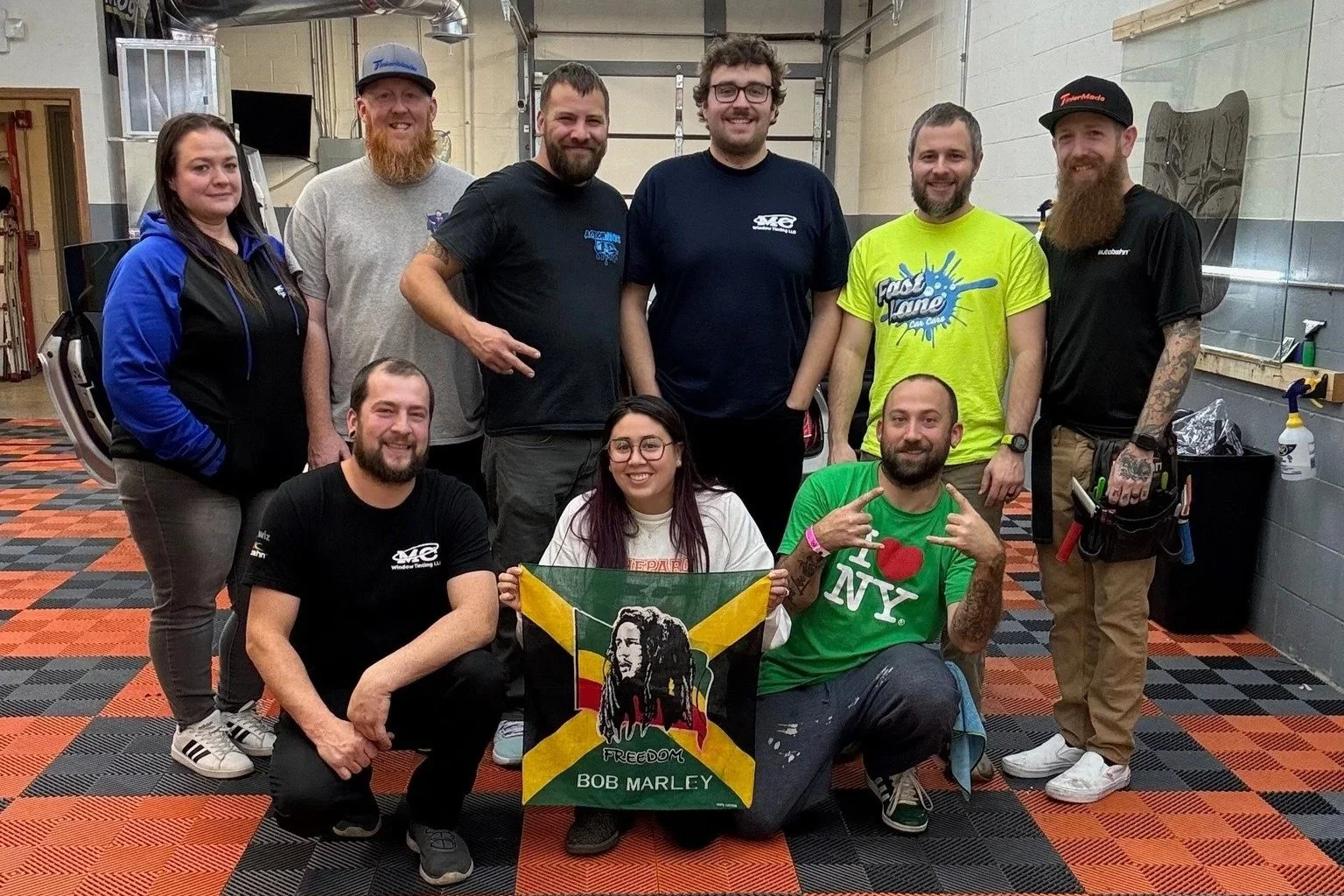 Group of ten people posing together indoors, some standing and some kneeling, with a fabric flag featuring Bob Marley's portrait and the words "Freedom Bob Marley" in front. The background shows a garage or workshop setting with tools and equipment.