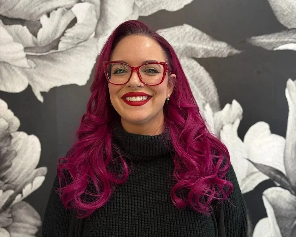 A woman with bright pink, curly hair, wearing red glasses, red lipstick, a black turtleneck sweater, and earrings, smiling in front of a black-and-white floral wallpaper background.