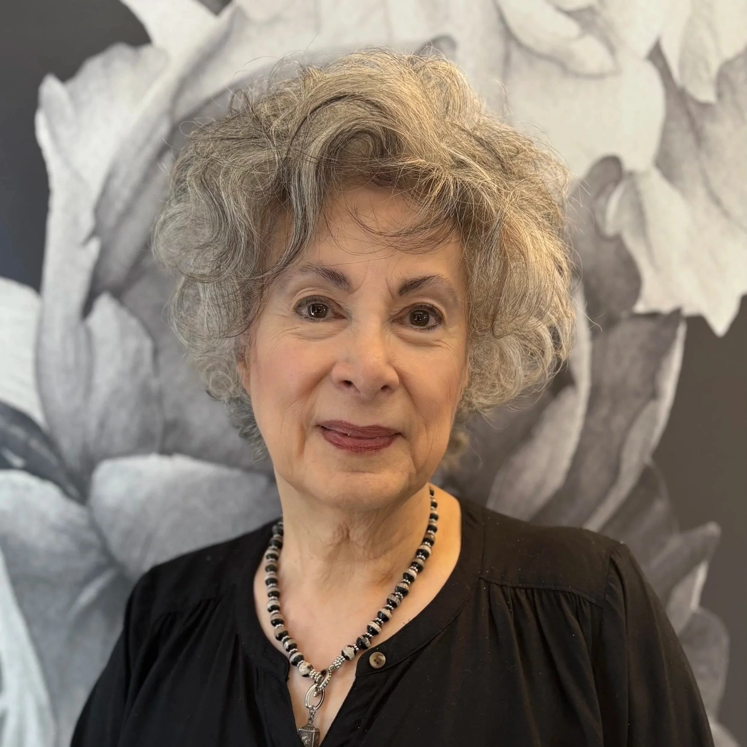 An elderly woman with grey, curly hair and wearing a black top stands in front of a large, black-and-white floral artwork. She is smiling and wearing a beaded necklace.